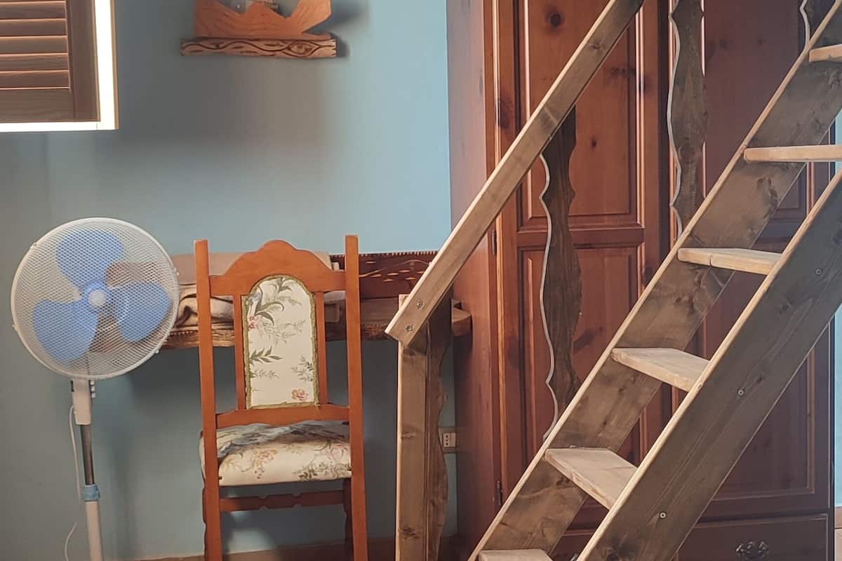 A room with a wooden staircase, a wooden chair with floral upholstery, and a standing fan.