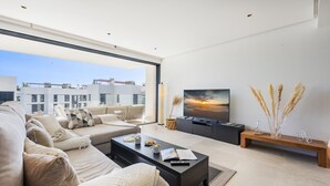 A modern living room with a large window, a sofa set, a coffee table, and a TV. - Penthouse  Apt Colomer by JS Villas (Port de Pollença)