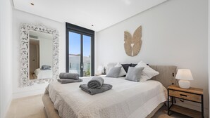 A bedroom with a large bed, a nightstand with a lamp, a mirror with an ornate frame, and a wall-mounted decorative piece. - Penthouse  Apt Colomer by JS Villas (Port de Pollença)