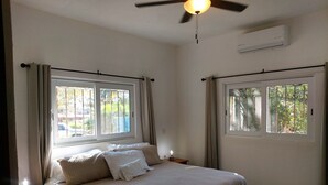 Une chambre à coucher comprenant un lit, une table de chevet, un ventilateur de plafond et deux fenêtres avec des rideaux.