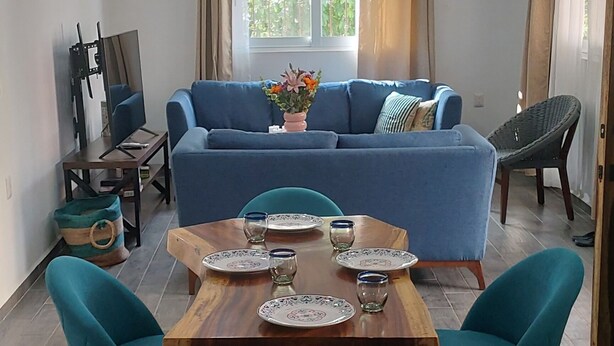 Un coin repas avec une table en bois dressée pour quatre personnes, entourée de chaises turquoise. Un canapé bleu est placé contre le mur, et un ventilateur de plafond est visible au-dessus.