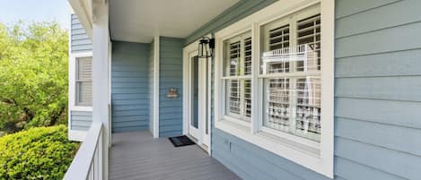 Une véranda avec un plancher en bois et des balustrades blanches, une pancarte d’accueil et une fenêtre ornée d’une grille décorative.