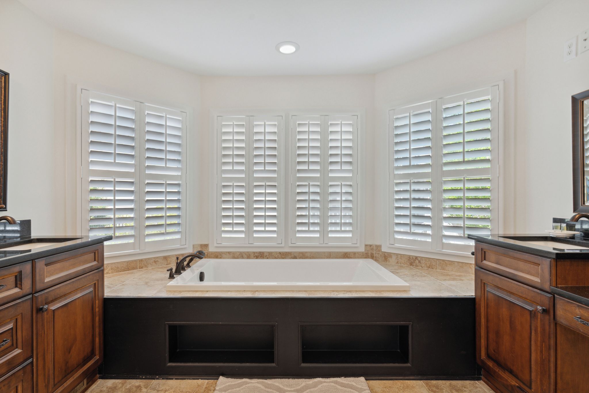 A bathroom with a freestanding bathtub, wooden cabinetry, and large windows with blinds.