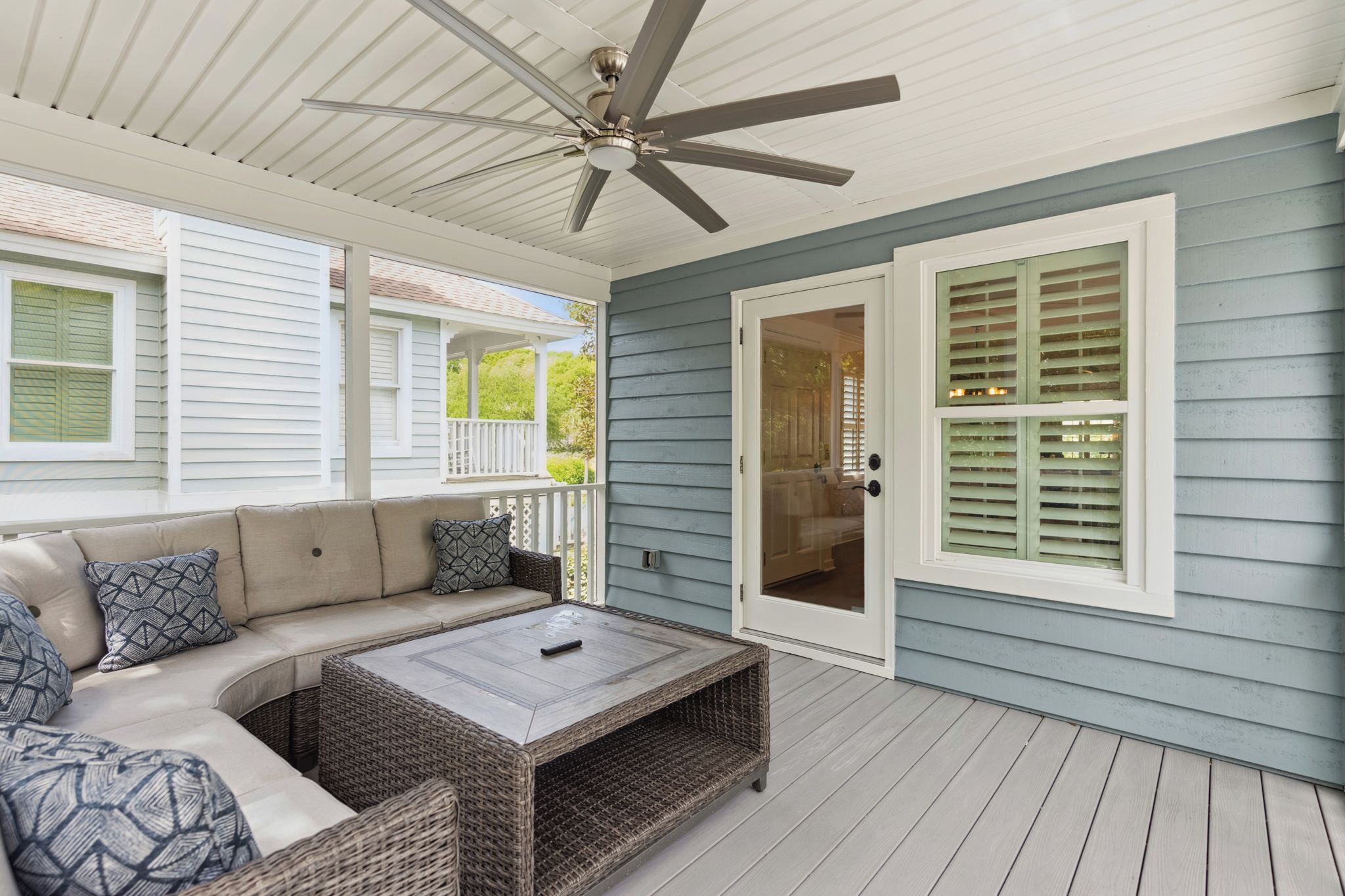 A covered porch with a ceiling fan, a cushioned seating area with a coffee table, and a door with green shutters.