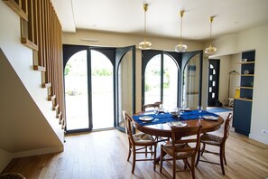 Un comedor con una mesa de madera, sillas y un camino de mesa azul. Se observan ventanas grandes con arcos y una escalera con barandillas de madera.