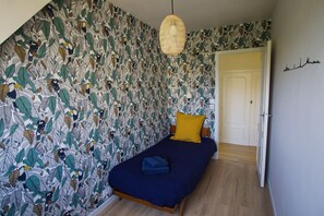 Un dormitorio con cama, almohada amarilla, manta azul y pared con estampado de hojas.