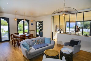 A modern living room with a grey sofa, a dining table with chairs, and a kitchen area visible through a glass door.