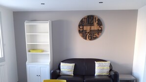 Een moderne woonkamer met een ronde glazen tafel, gele stoelen, een zwarte bank met gekleurde kussens en een houten wandklok.