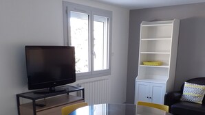 Een moderne woonkamer met een ronde glazen tafel, gele stoelen, een flatscreen televisie op een houten standaard, een witte boekenkast, en een grijze bank met gestreepte kussens.