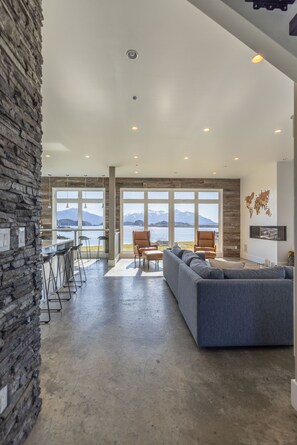 A spacious living area with a large window overlooking a lake and mountains, a stone wall, and a modern bar area.