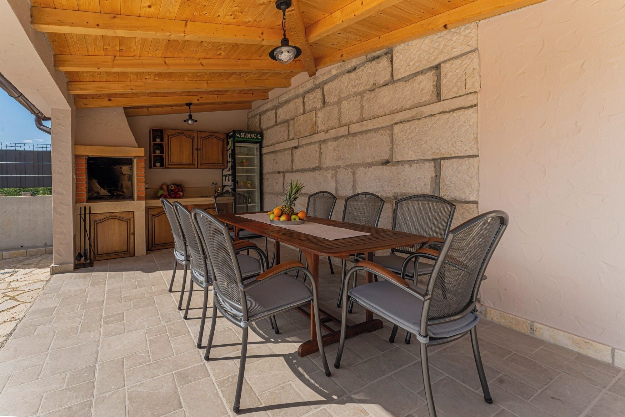 An outdoor dining area with a wooden table and metal chairs, a stone wall, and a stone fireplace.