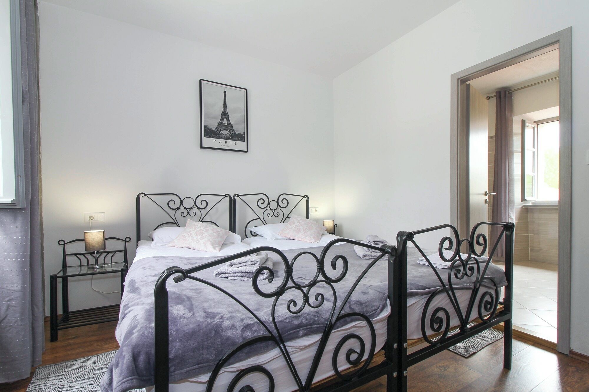Ein Schlafzimmer mit einem Metallbettgestell, einem Einzelbett mit weißer Bettwäsche, einem kleinen Nachttisch mit einer Lampe, einem gerahmten Bild des Eiffelturms und einer Tür zu einem anderen Raum.