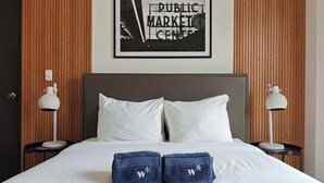 Una cama bien tendida con sábanas blancas, dos almohadas y dos toallas dobladas con un logotipo en una habitación de hotel.
