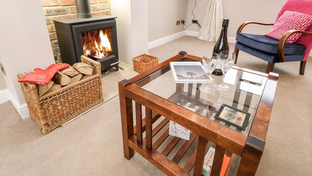 A cozy living room with a fireplace, a glass coffee table with books and glasses, a wicker basket, and a blue chair with a pink cushion. - Willows (Corbridge)