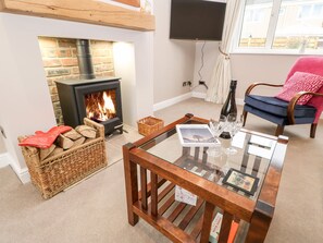A cozy living room with a fireplace, a glass coffee table with books and glasses, a wicker basket, and a blue chair with a pink cushion. - Willows (Corbridge)