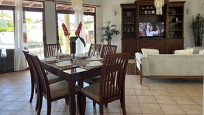 Uma sala de jantar com uma mesa de madeira posta para seis pessoas, rodeada por cadeiras, e uma sala de estar com sofá e televisão.