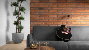 Une pièce avec un mur en briques, un canapé gris, une guitare noire, une plante en pot et une table sur laquelle se trouvent deux verres et un bol de chocolats.