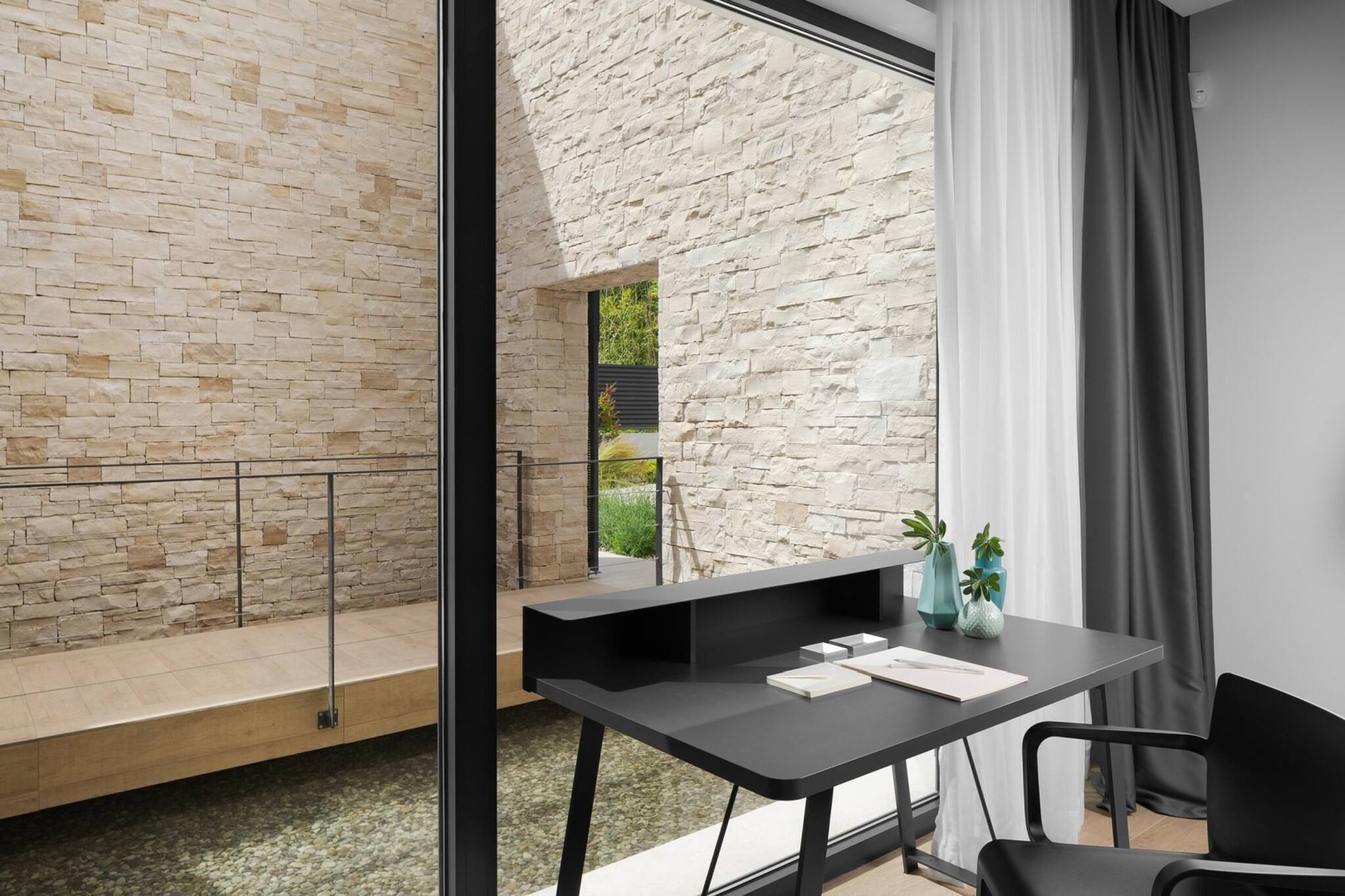 A modern room with a stone wall, a glass door leading to an outdoor area, a black desk with a chair, and a window with curtains.