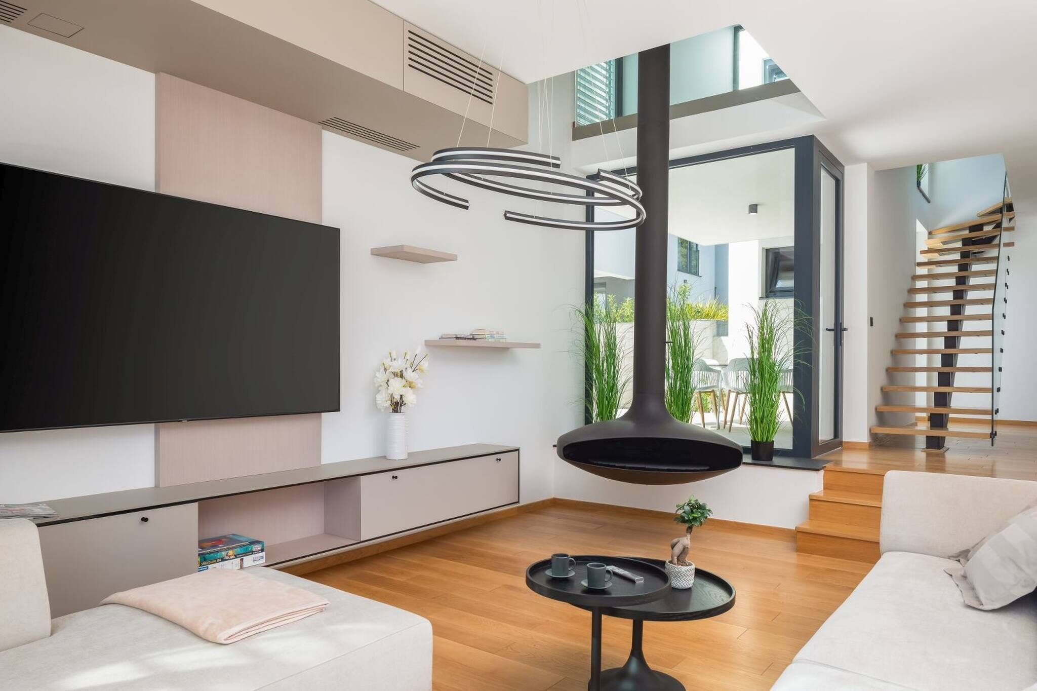 Een moderne woonkamer met een grote flatscreen televisie, een hanglamp, een trap en uitzicht op groen buiten.
