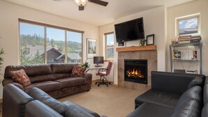 A living room with a fireplace, leather sofas, a TV, and large windows offering a view of the outdoors.