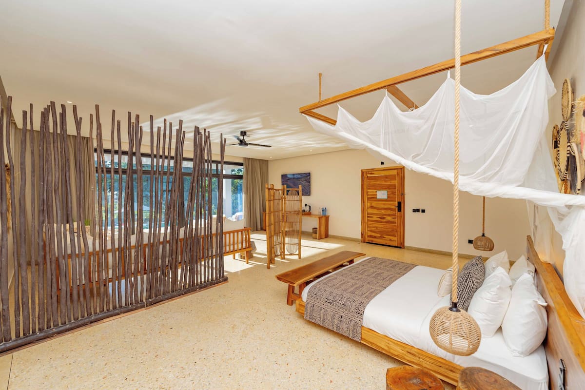 Un dormitorio amplio con una cama grande, un banco de madera, un dosel colgante sobre la cama y vistas al exterior.