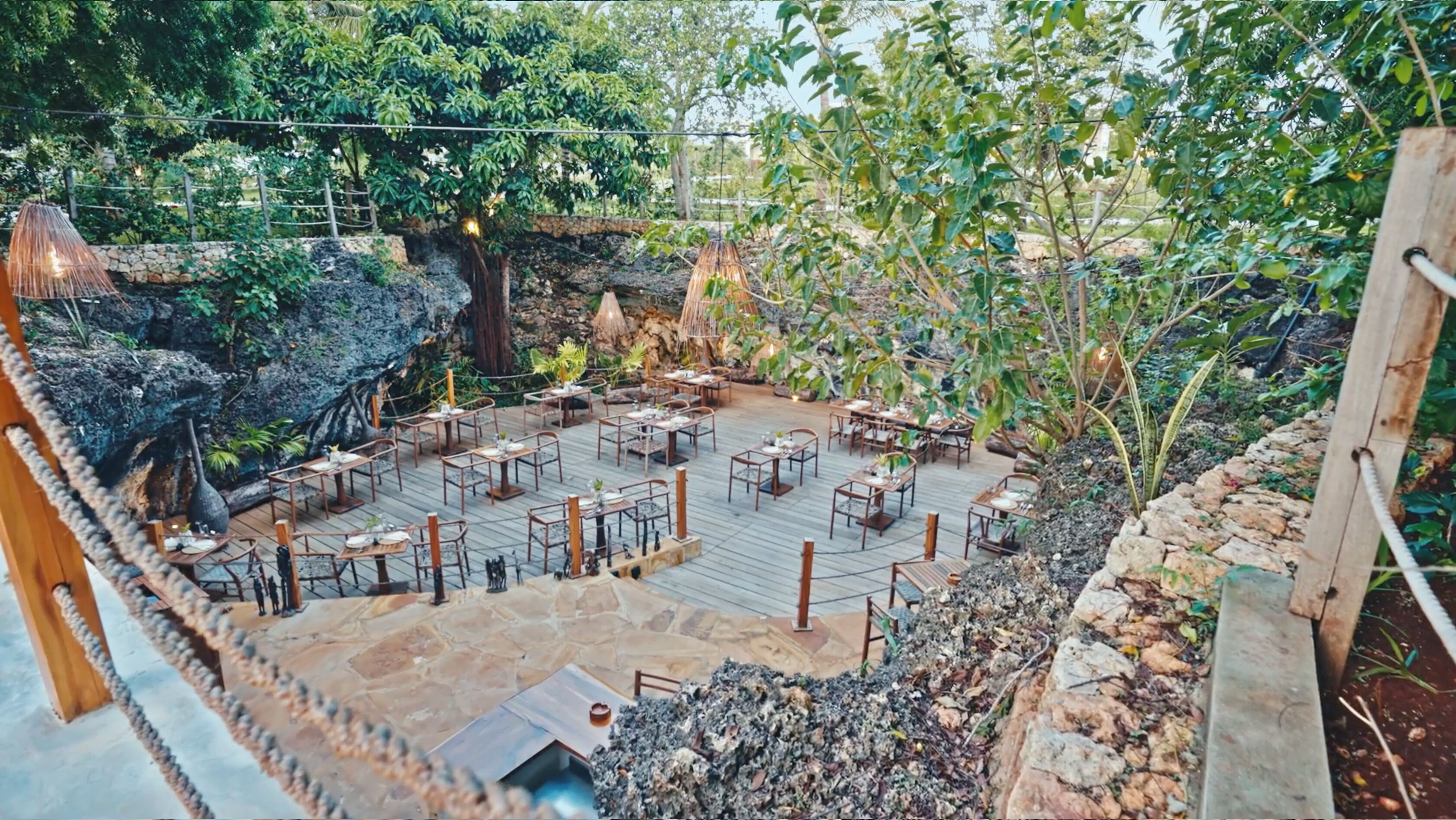 An outdoor dining area with tables and chairs, surrounded by greenery and a rocky landscape.