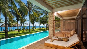 A poolside area with wooden loungers, a stone pillar, and a view of the ocean and palm trees.