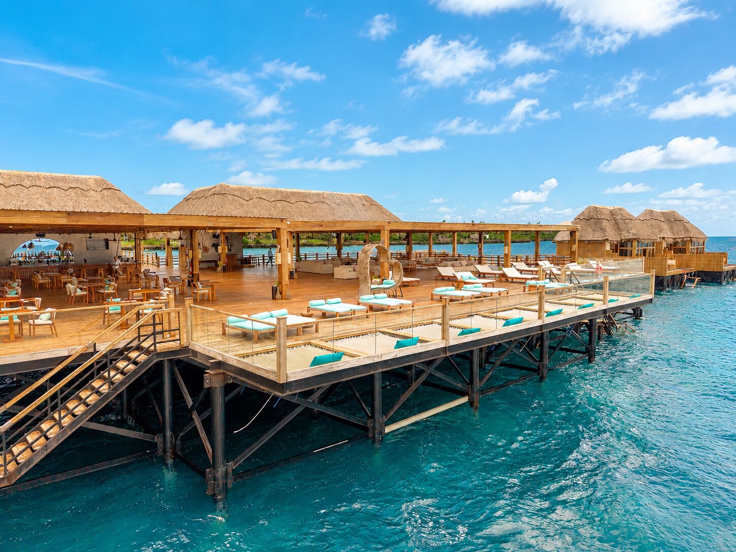 An overwater bungalows with wooden deck seating and thatched roofs, connected by a wooden walkway over clear blue water.