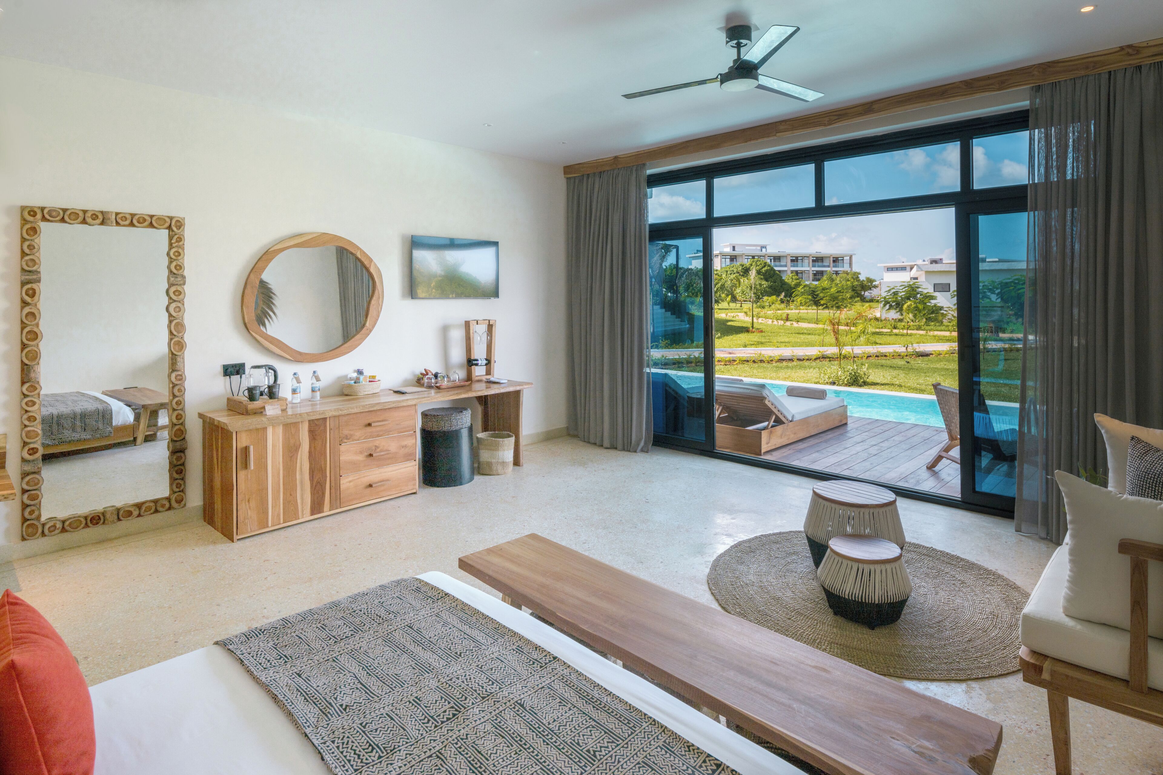 A modern hotel room with a large bed, a wooden dresser, a desk, a large mirror, and a view of a swimming pool and greenery outside.