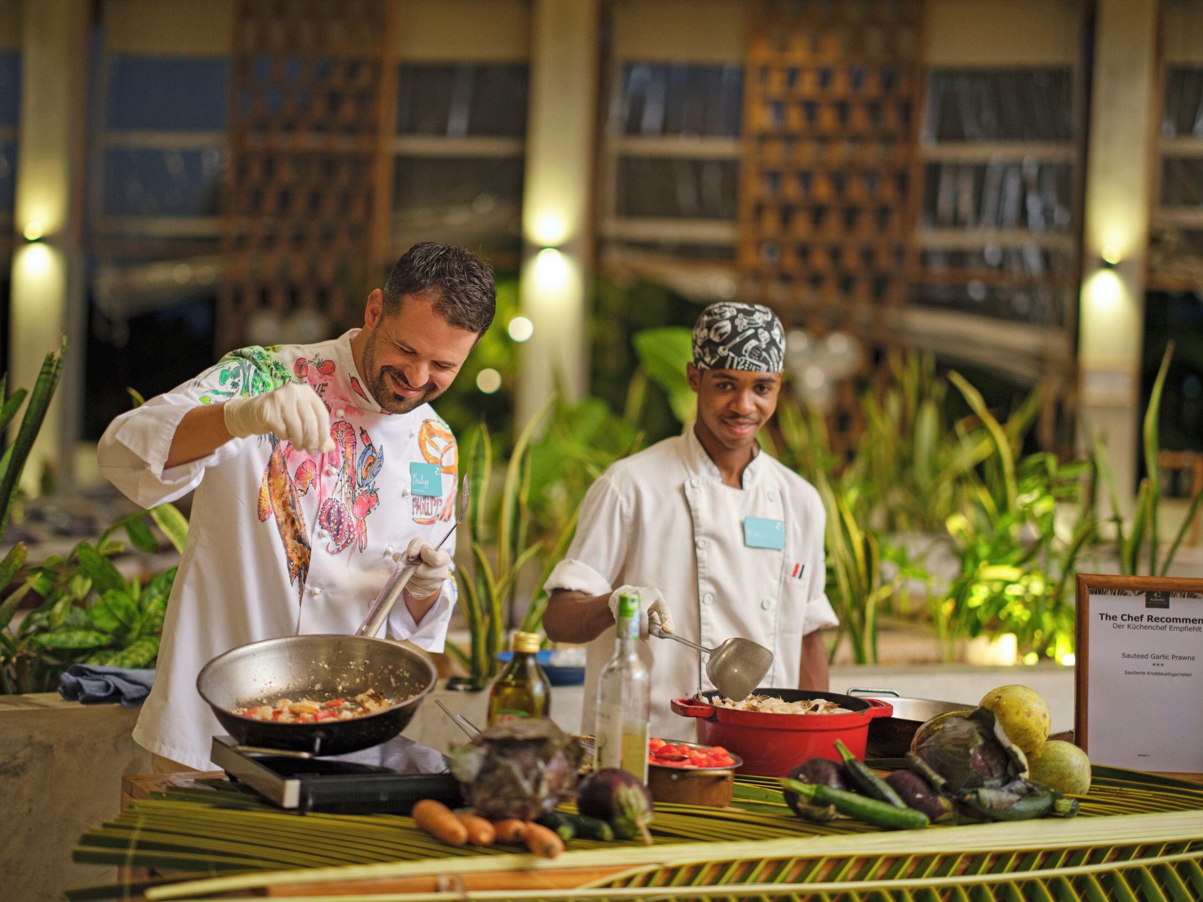 Two chefs preparing food in a well-lit indoor setting with a variety of vegetables and cooking utensils on the table.