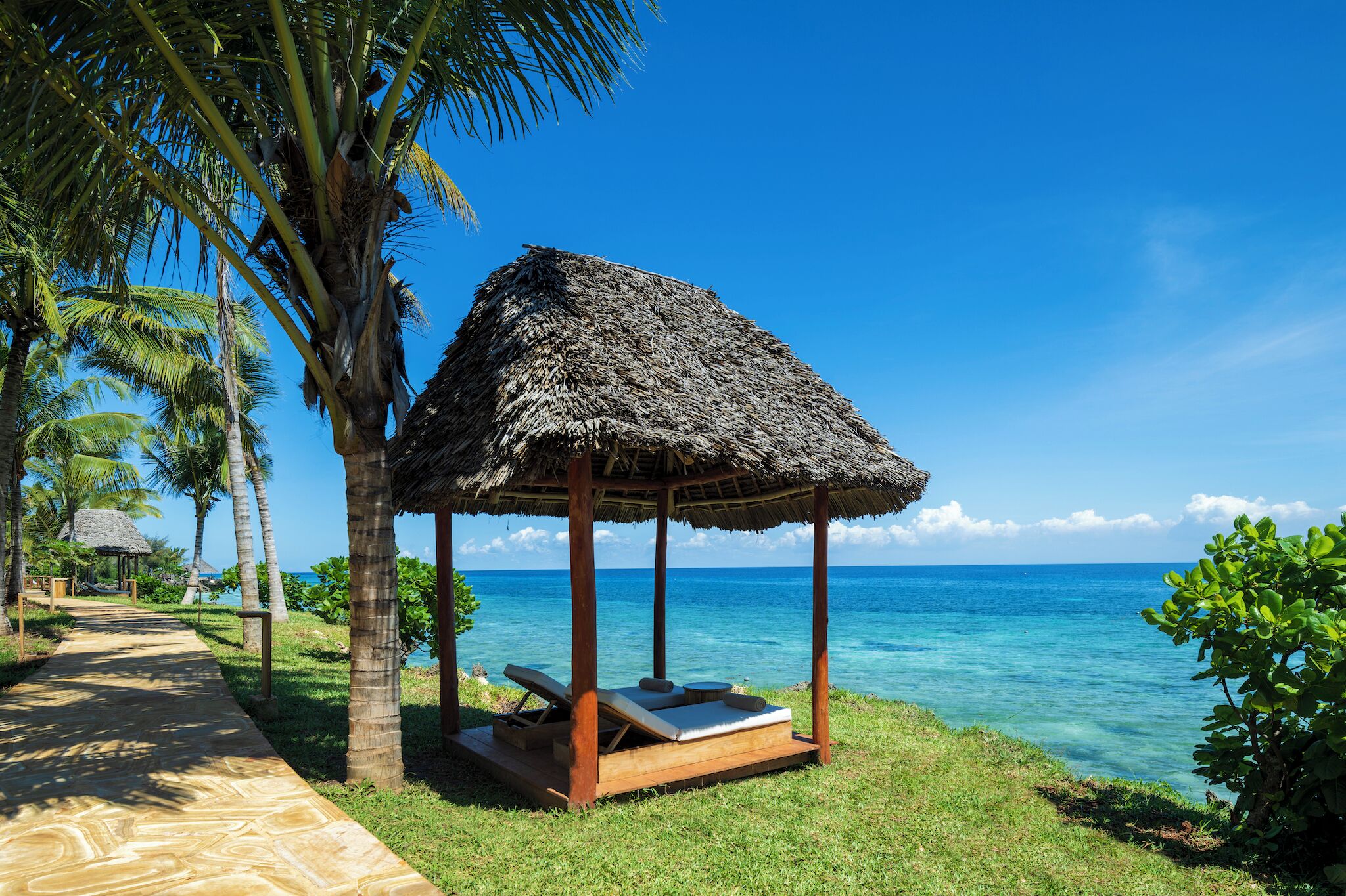 A thatched-roof gazebo with a bench and a small table, surrounded by palm trees and a clear view of the ocean.