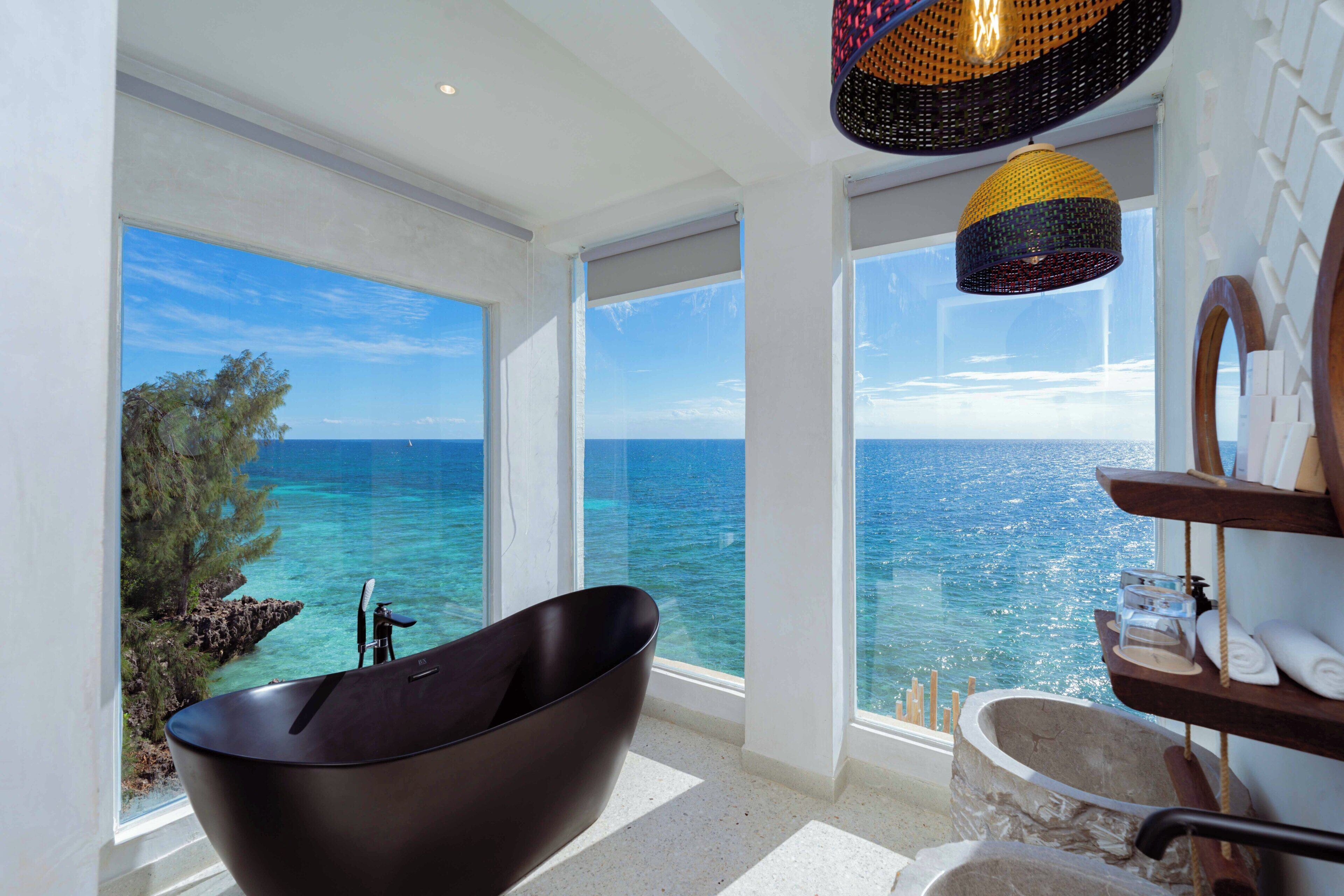 Um banheiro moderno com uma banheira livre, amplas janelas com vista para o mar e um lustre de teto singular.