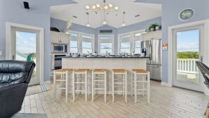Una cocina moderna con armarios blancos, una isla central con taburetes de bar, electrodomésticos de acero inoxidable y grandes ventanas con vistas al exterior.
