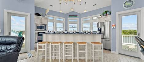 A modern kitchen with white cabinetry, a central island with bar stools, stainless steel appliances, and large windows offering a view of the outdoors.