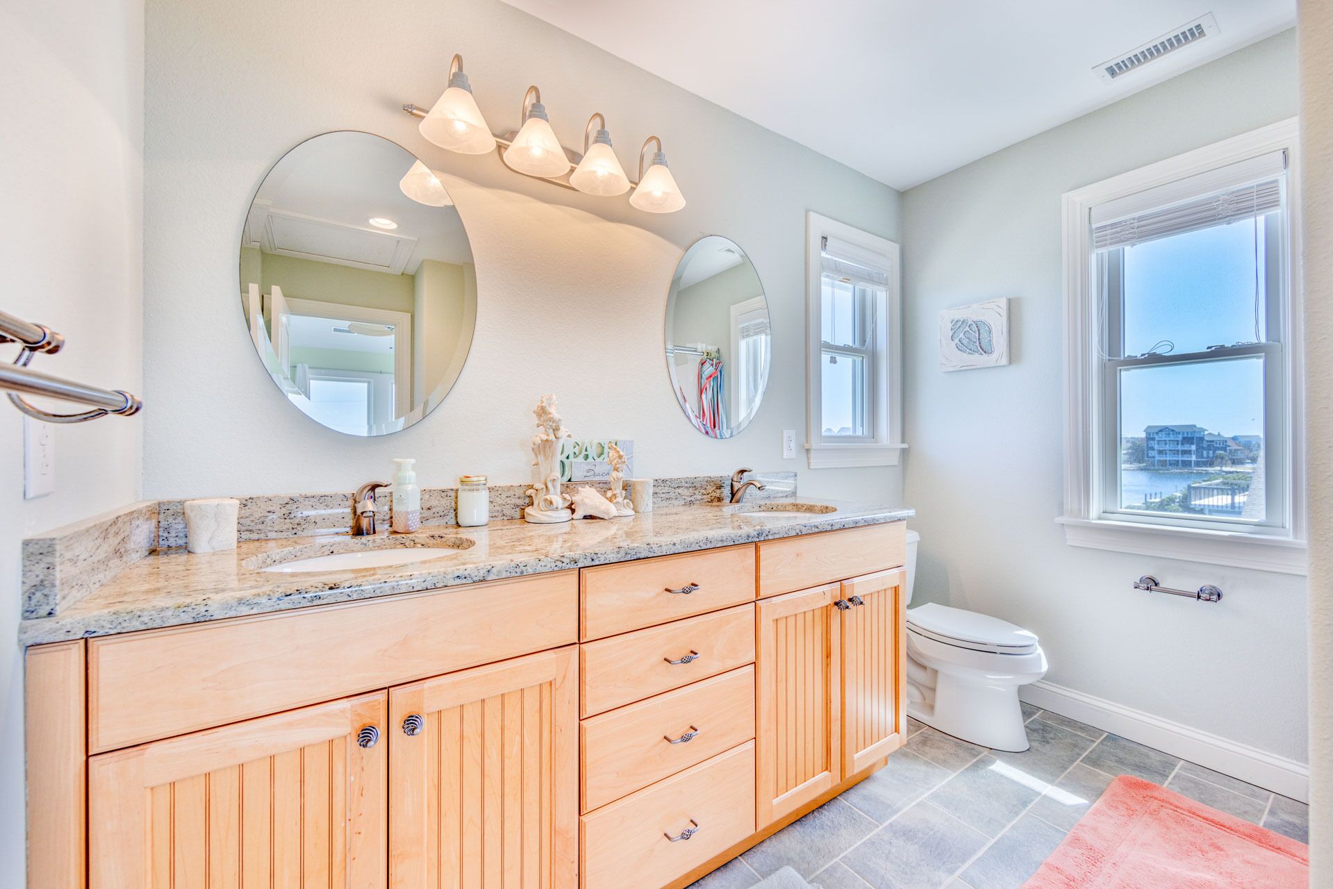 A bathroom with a granite countertop, wooden cabinets, a white toilet, and a large window with a view.