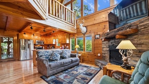 A spacious living area with a wooden floor, a sofa, a green armchair, a fireplace, and a kitchen in the background.