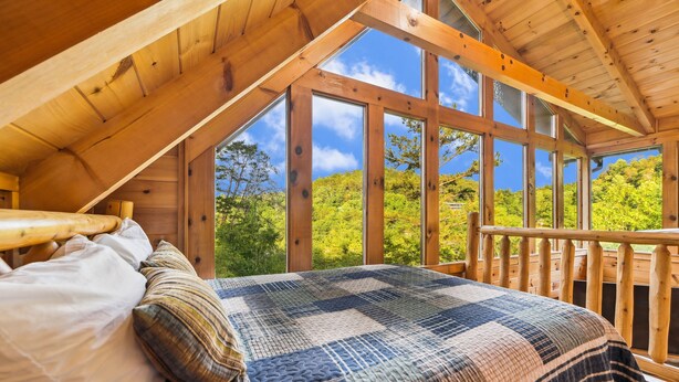 A bedroom with a large bed, wooden ceiling beams, and a view of a forest through large windows.