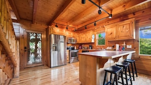 A wooden cabin interior with a kitchen featuring stainless steel appliances, a large island with bar stools, and a view of greenery outside.