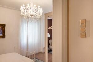 Habitación de hotel con candelabro, cama, puerta con placa de número y un cuadro en la pared.