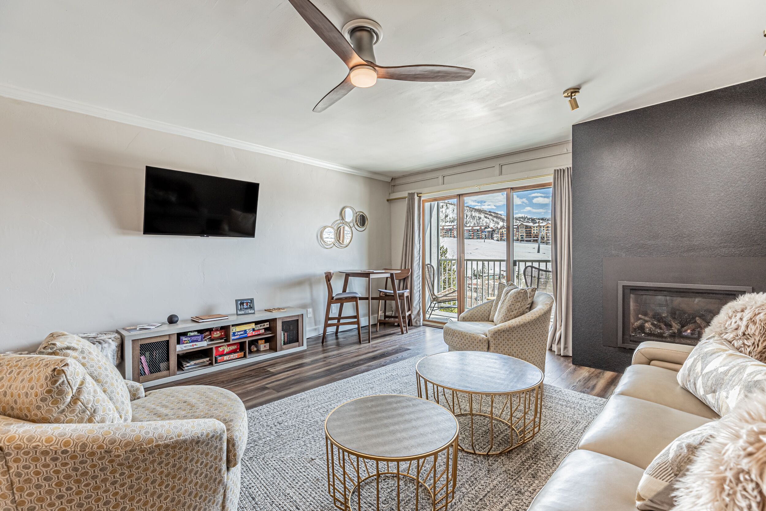 A living room with a fireplace, a ceiling fan, a TV mounted on the wall, and a view of the outdoors.