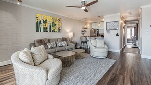 Ein modernes Wohnzimmer mit einem grauen Sofa, einem gemusterten Sessel und einem runden Couchtisch. Eine Deckenleuchte und Einbauleuchten sind zu sehen.