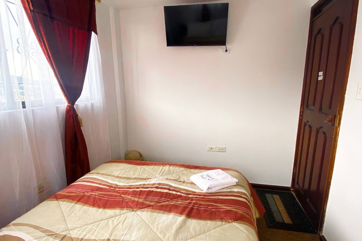 A hotel room with a bed, a television on the wall, a door, and a window with curtains.