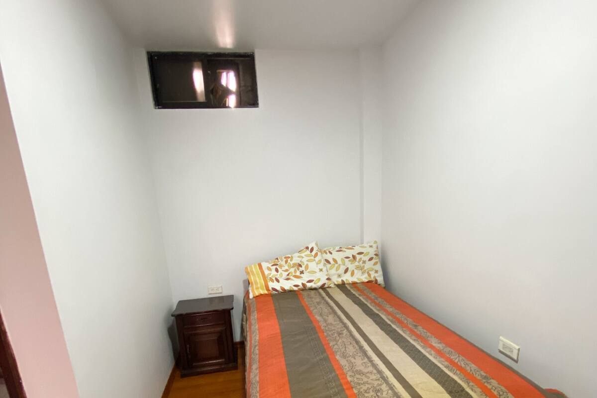 A small, single-bed room with a wooden floor, a bedside table, and a wall-mounted light fixture.