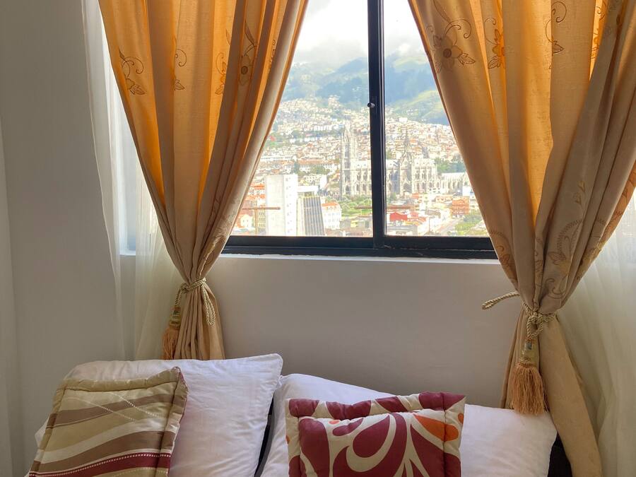 A bed with patterned pillows and a towel, curtains tied back, and a cityscape view through a window.