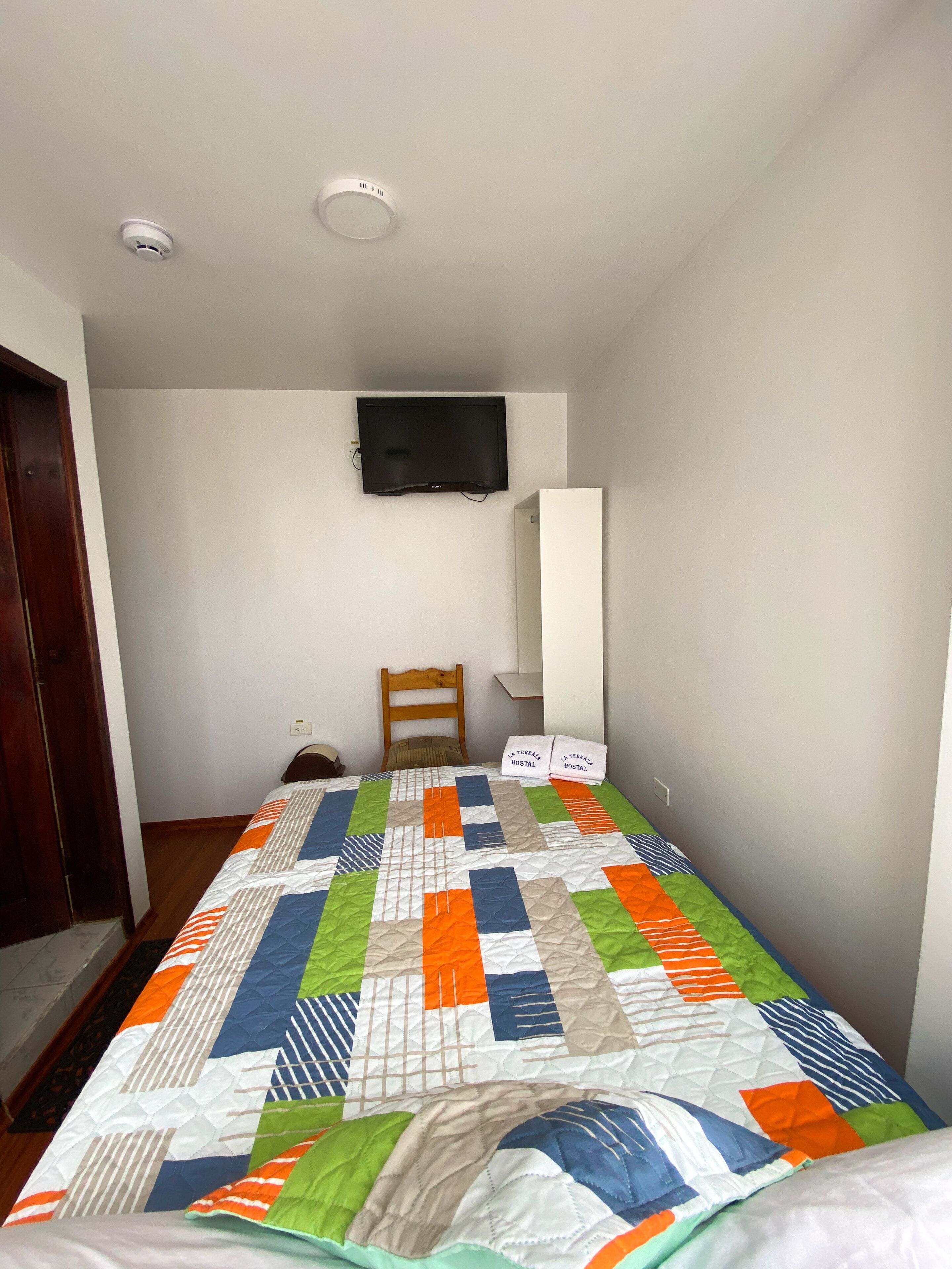 A small, simple room with a bed, a wooden chair, a flat-screen TV mounted on the wall, and a white wardrobe.