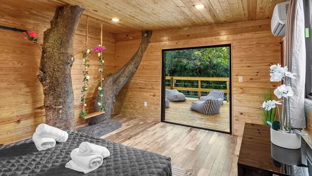A wooden cabin room with a bed, towels, a hanging plant, and a view of an outdoor deck.