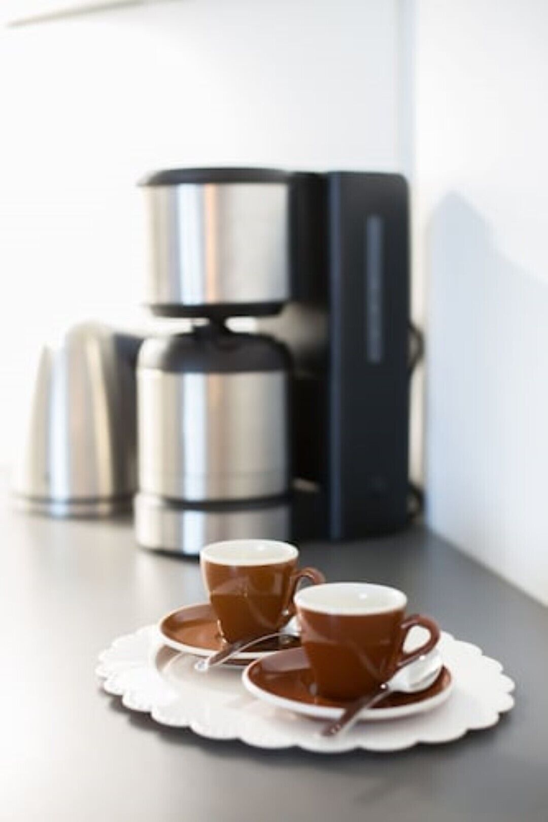 Twee koffiekopjes op een witte tafelkleed met een lepel, een koffiezetapparaat en een stapel koffiecontainers op een tafel.