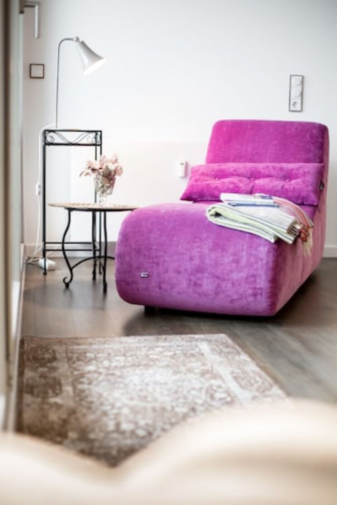 Un fauteuil violet avec un magazine plié, une petite table sur laquelle est posé un vase de fleurs et une lampe sur pied.