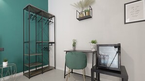A room with a teal accent wall, a black metal shelving unit with clothes, a green chair, a small table with a potted plant, and a framed quote on the wall. - Chianu Palì guest rooms (Agrigento)