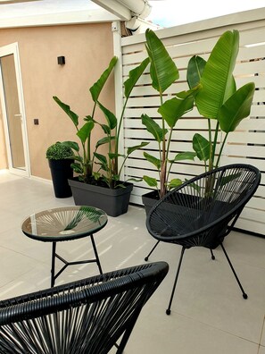 Un espacio de patio con dos sillas de mimbre negro, una mesa de vidrio y varias plantas en macetas, incluyendo una planta de hoja de plátano grande.